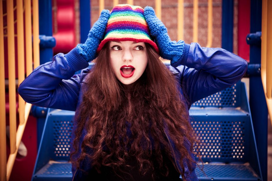 A picture of Kate Sloan, a white woman with long, wavy dark hair. She has her hands on the side of her head and her mouth open in a 'surprised' expression. She is wearing a blue jacket, blue gloves and a rainbow striped knitted hat.