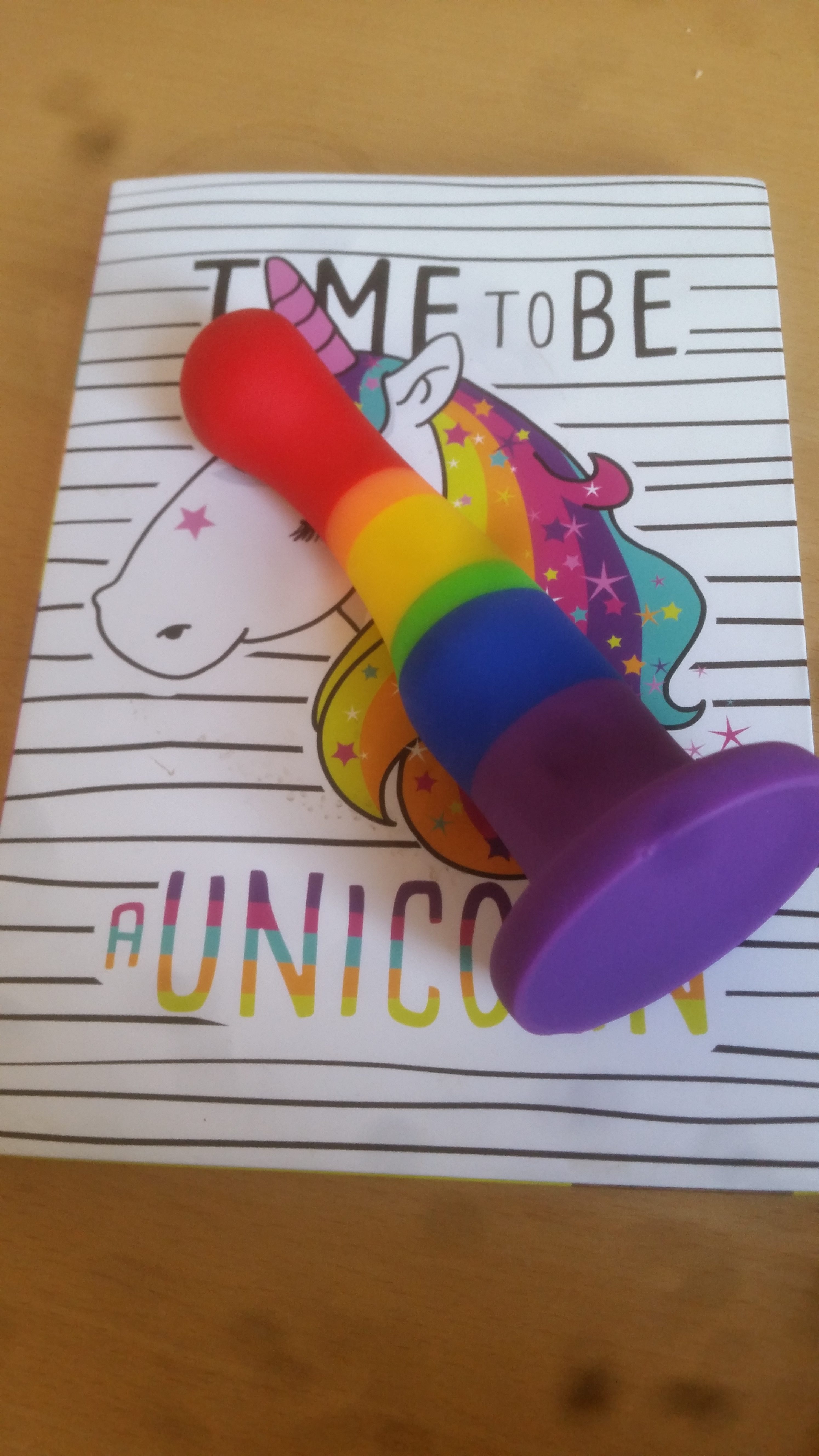 A rainbow dildo lying on a white notebook with a unicorn on the cover.