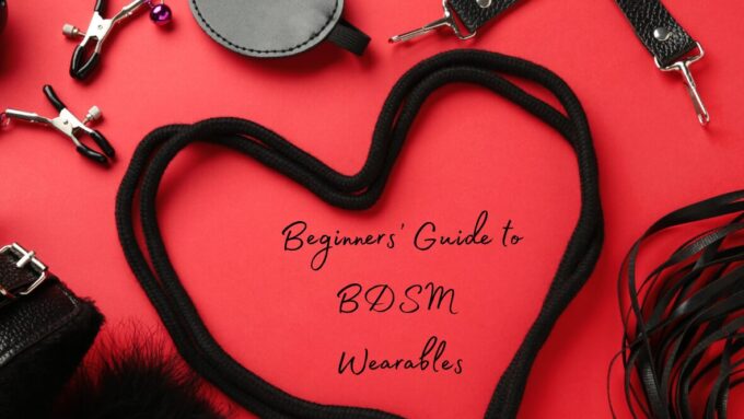 Several BDSM items including leather straps, nipple clamps, and a blindfold on a red background. A length of black rope forms a heart. Text in the middle of the heart reads: A Beginners' Guide to BDSM wearables